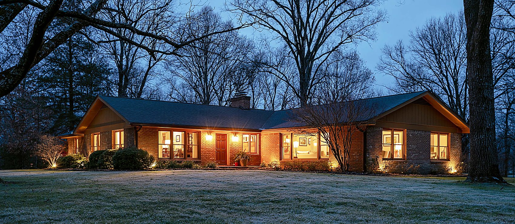 A brick ranch house glowing with warm light in every window at winter twilight, with landscape lighting along the frosted front lawn