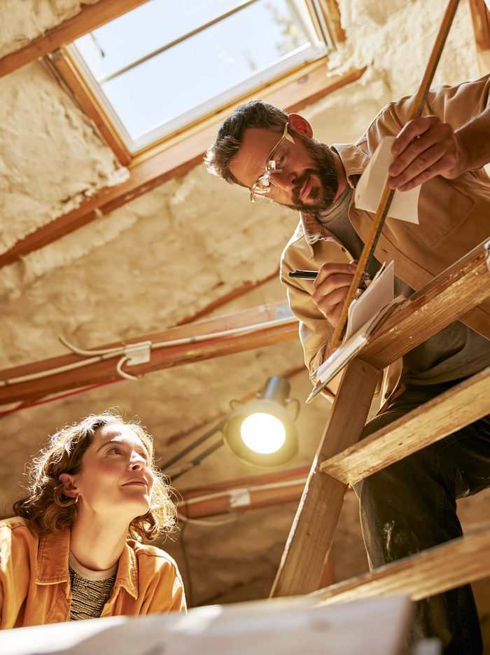 An estimator measuring insulation depth at the top of an attic ladder while the homeowner watches from below