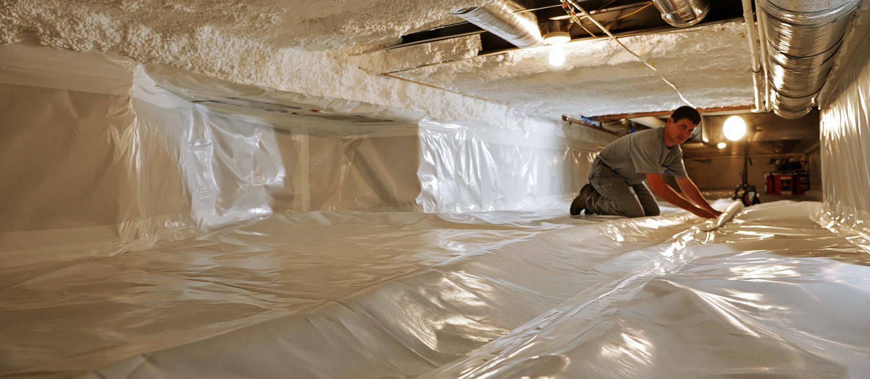 Installer smoothing a white vapor barrier across a sealed crawl space floor, with foam-coated joists and ductwork overhead