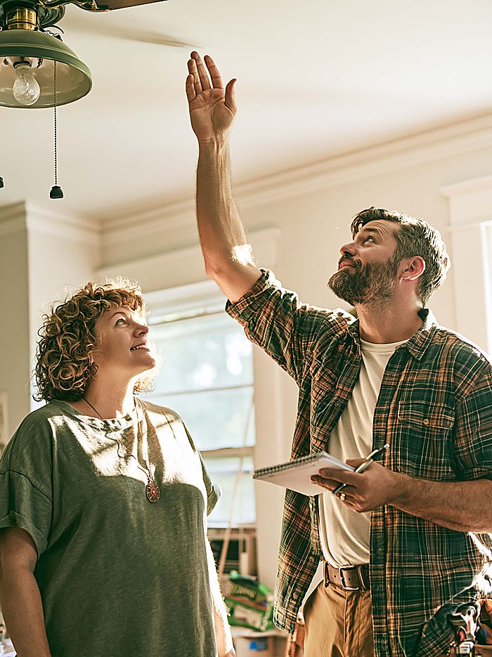 An estimator reaching up to feel for a draft at a ceiling light fixture while the homeowner looks on, notepad in hand