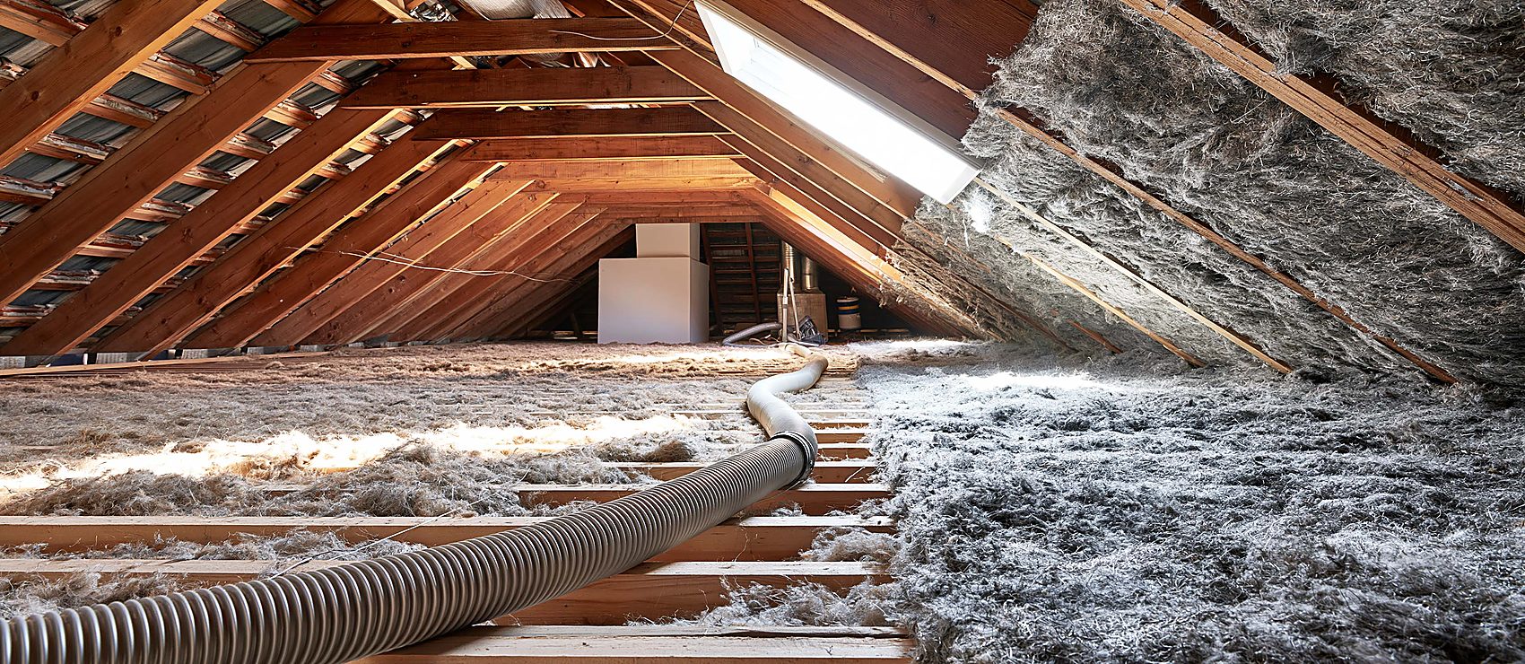 Attic interior mid-cleanout with a flexible hose feeding insulation into the bay on one side while the wood roof decking sits bare on the other