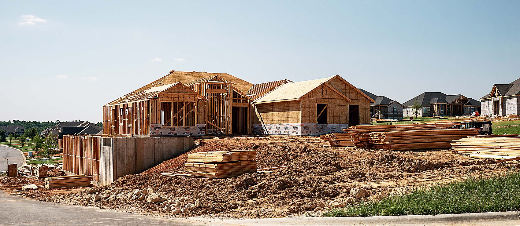 Two newly framed houses on a subdivision lot, one set on a tall concrete foundation wall on the downhill side, with stacked lumber and graded dirt out front